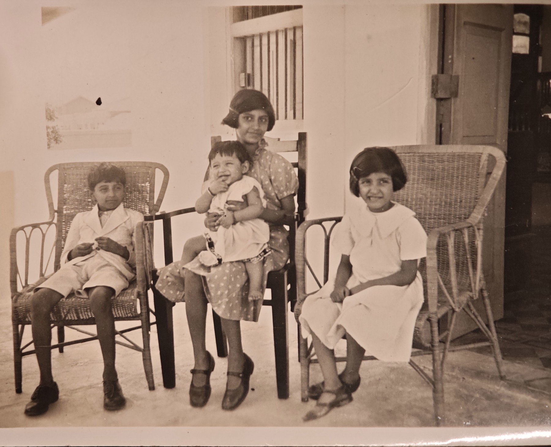 The four Dubash children on cane chairs on a veranda in Karachi, the eldest girl holding the youngest on her lap.