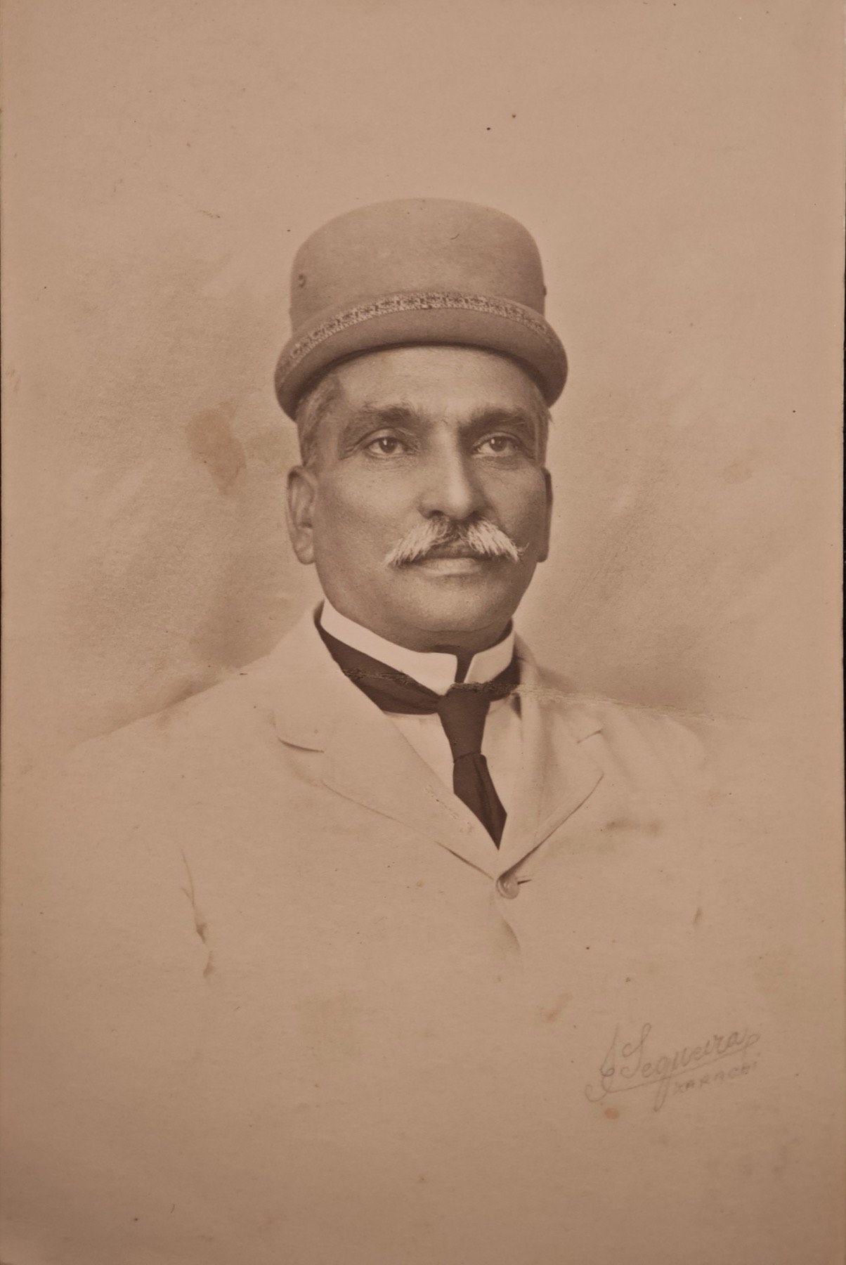 Sepia studio portrait of a man with a moustache, wearing a Parsi pagri, a pale jacket and a dark tie.