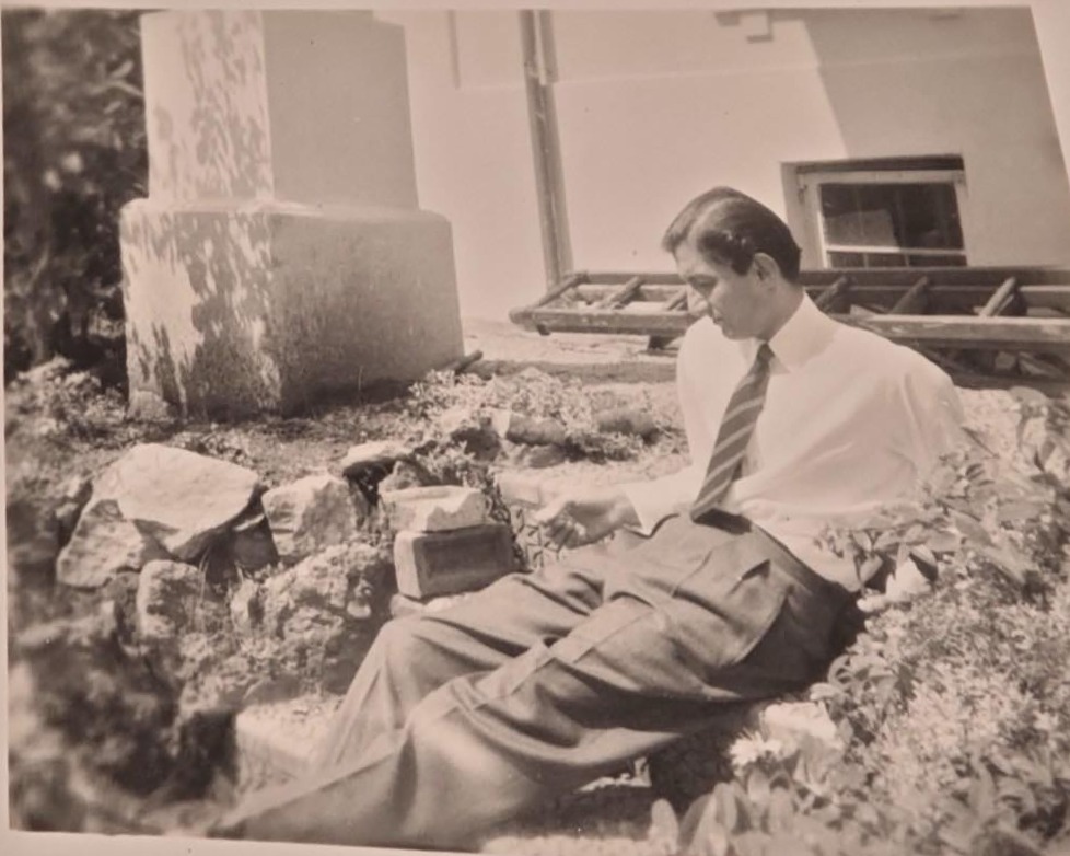 Sam in a shirt and tie, sitting back on a rockery in a garden in Paignton.
